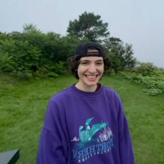 贝克微笑着，头戴反帽，身穿紫色运动衫，上面是绿色的雾蒙蒙的风景.